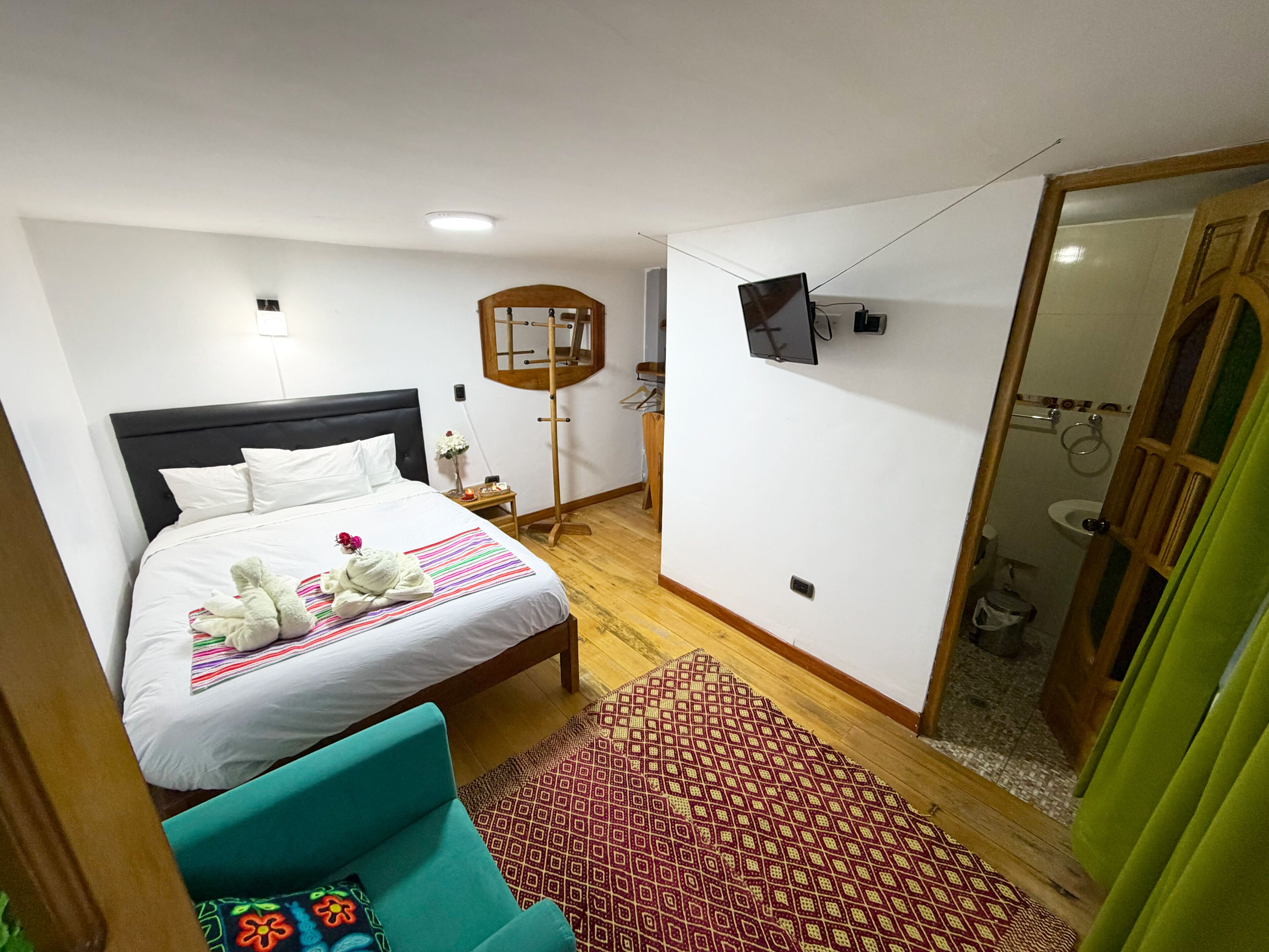 A hotel room with a bed, a small table, a mirror, a TV, and a bathroom visible through an open door.