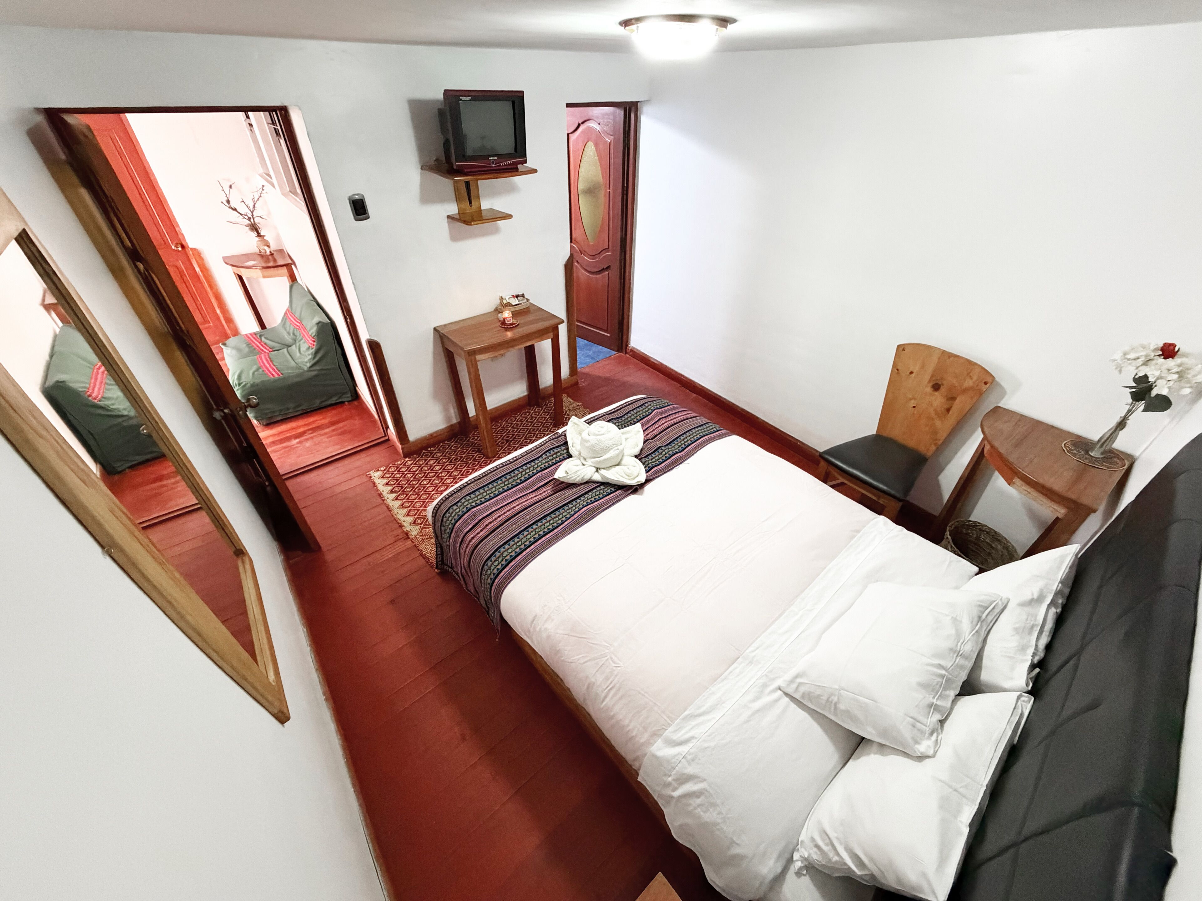 Una habitación de hotel con una cama, televisión, una mesita, una silla, un espejo y una puerta que da a otra habitación.