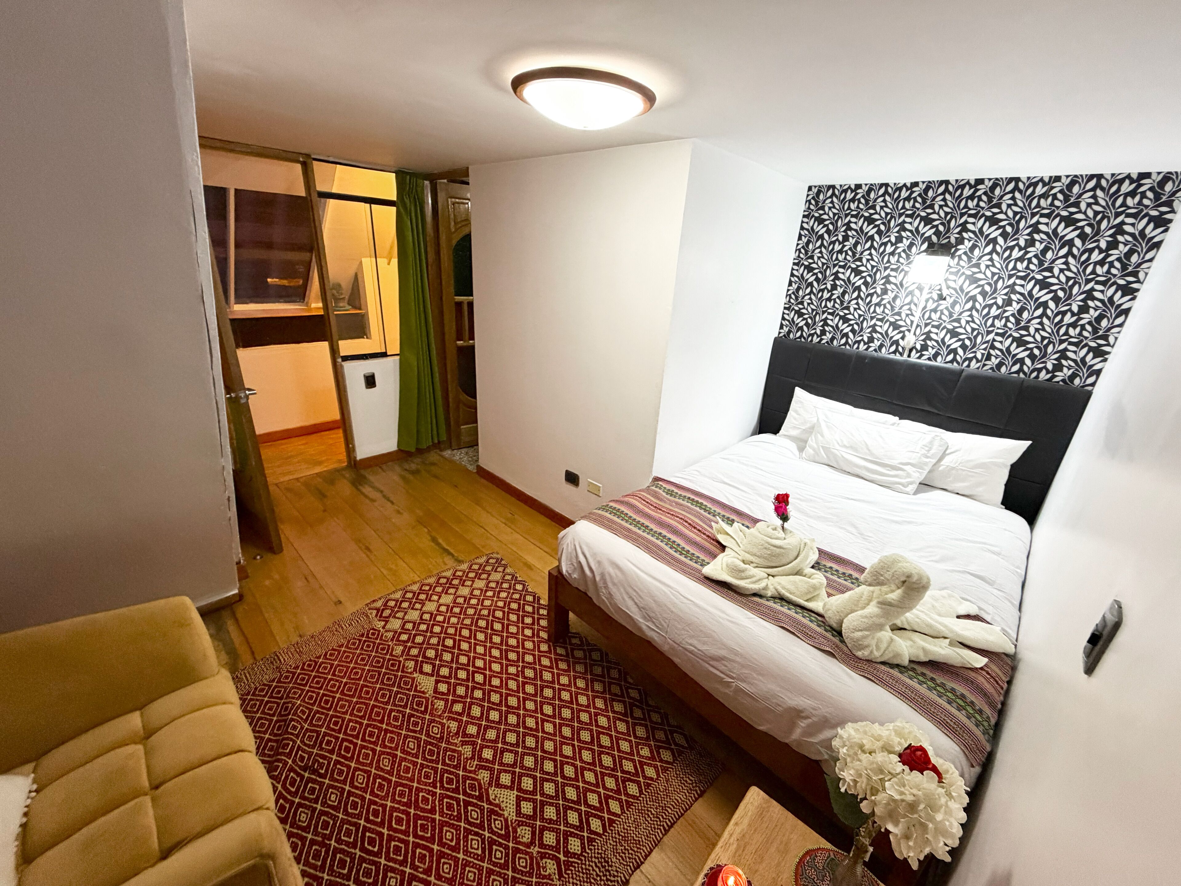 Una habitación de hotel con una cama, una mesita con una vela, una silla, un tapete estampado y una pared con un diseño de hojas.