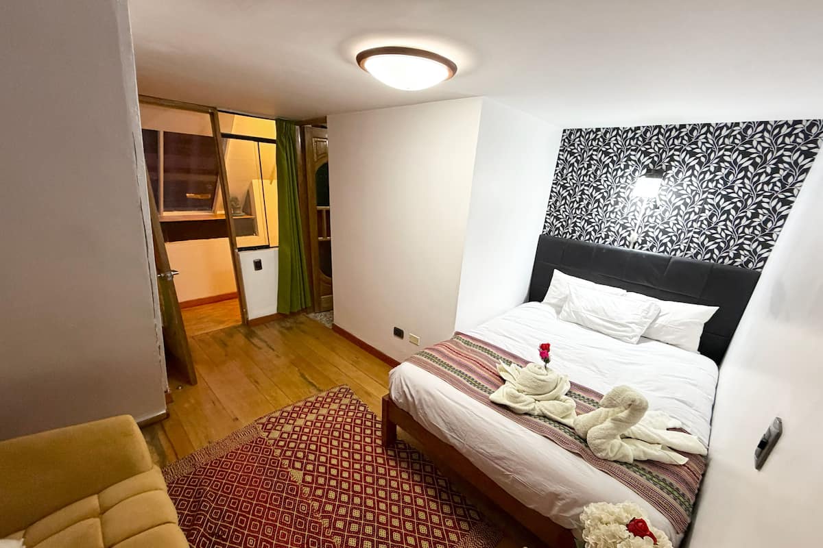 Una habitación de hotel con una cama, una mesita con una vela, una silla, un tapete estampado y una pared con un diseño de hojas.