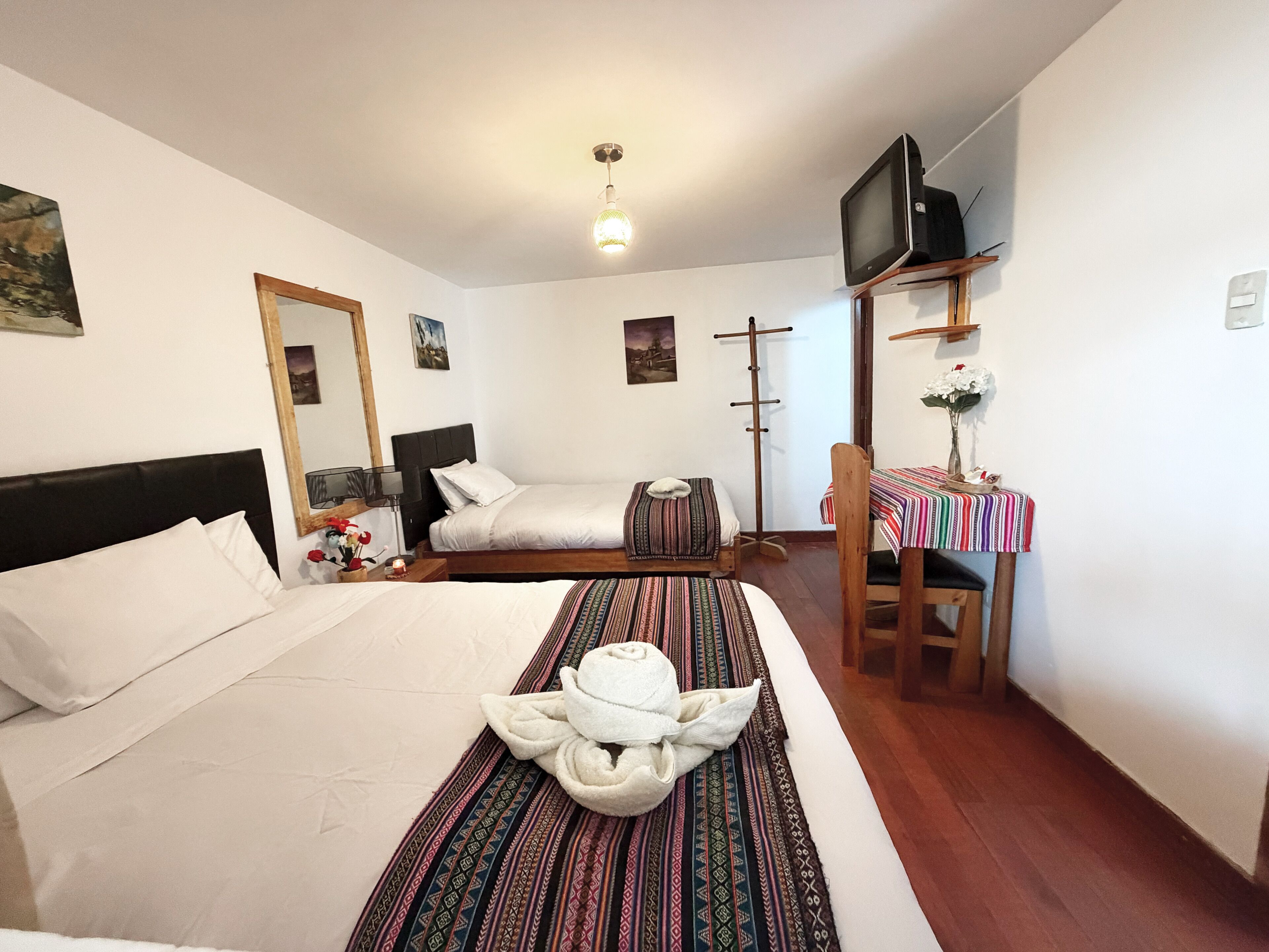 Una habitación de hotel con una cama, una mesita con televisión, una silla y un perchero.