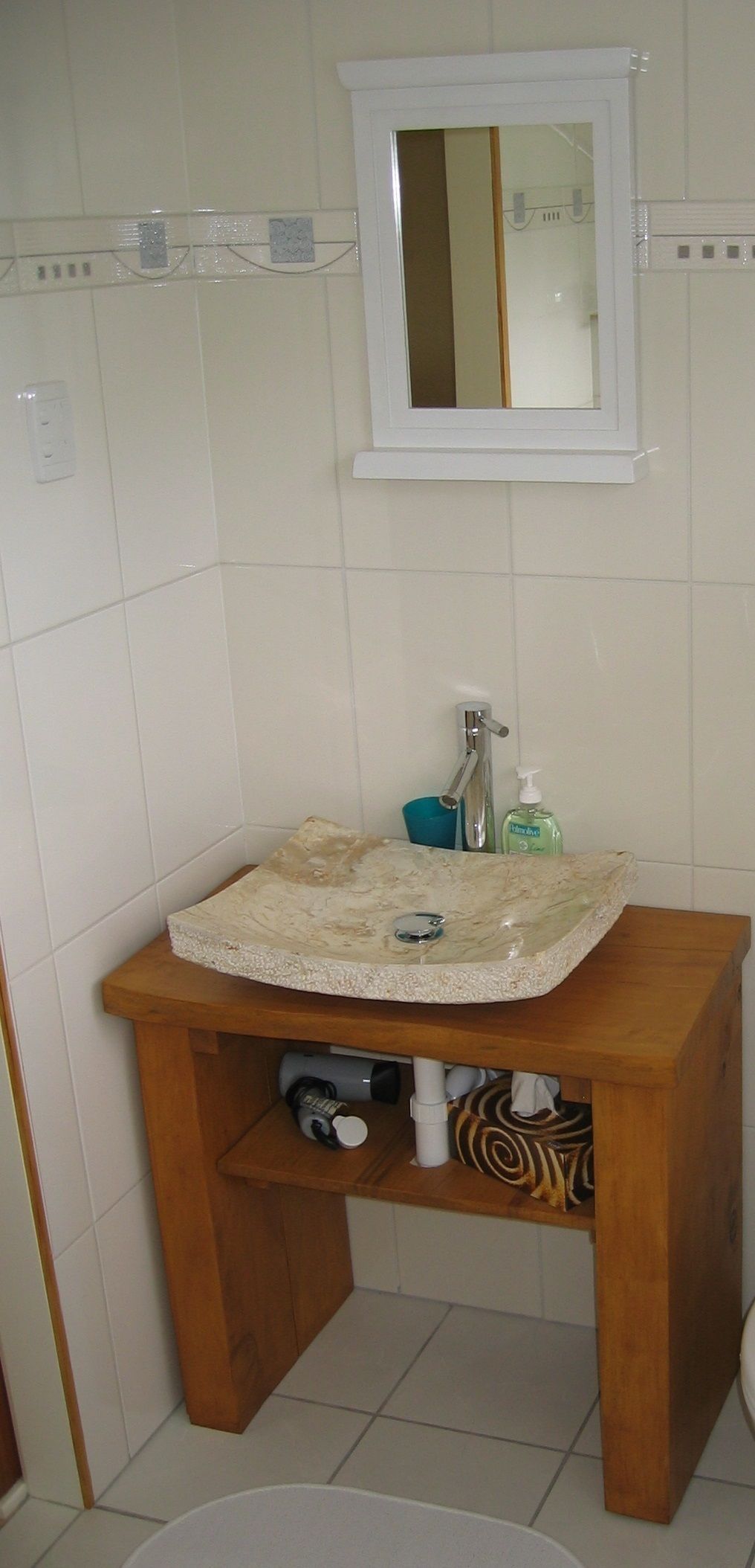 Une salle de bain avec un mur recouvert de carreaux blancs, un meuble-lavabo en bois, un miroir et une petite étagère au-dessus du miroir.