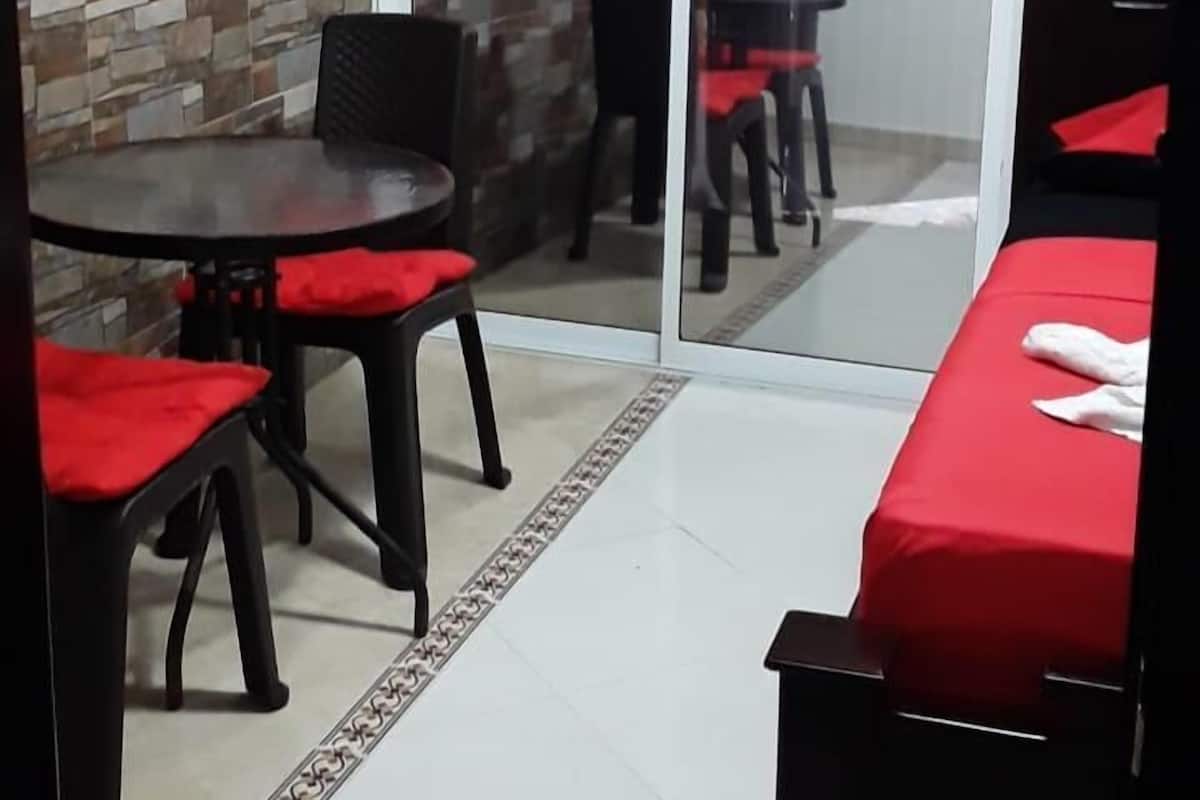 A room with a tiled floor, a glass door leading outside, a dining table with chairs, and a bed with a red cover.