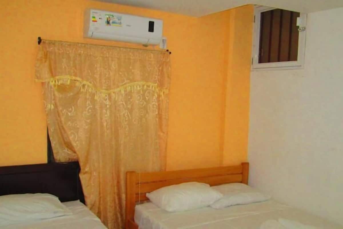 A room with two beds, a window with blinds, and an air conditioning unit on the wall.