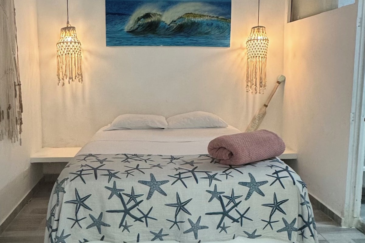 Un dormitorio con cama, dos lámparas colgantes, un cuadro de olas y una silla de madera.