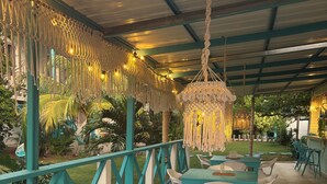 Un espace extérieur aménagé avec des tables turquoise et des chaises blanches, entouré d’une rambarde verte et agrémenté de décorations en macramé suspendues.