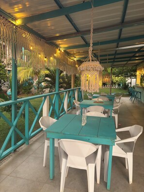 Een terras met turquoise tafels en witte stoelen, omgeven door een groene balustrade en macramé decoraties.