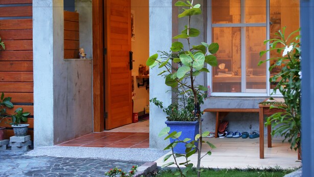 Entrada moderna de un edificio con puerta de madera, una maceta azul con un árbol pequeño y un banco con zapatos debajo.