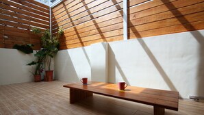 Un banco de madera con dos tazas rojas, una planta en maceta y un suelo de baldosas en una habitación soleada con listones de madera en la pared.