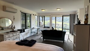 A hotel room with a large bed, a desk, a chair, and a sofa. There are large windows with a view of the countryside. - Mohua Park - Catlins Eco Accommodation (Tawanui)