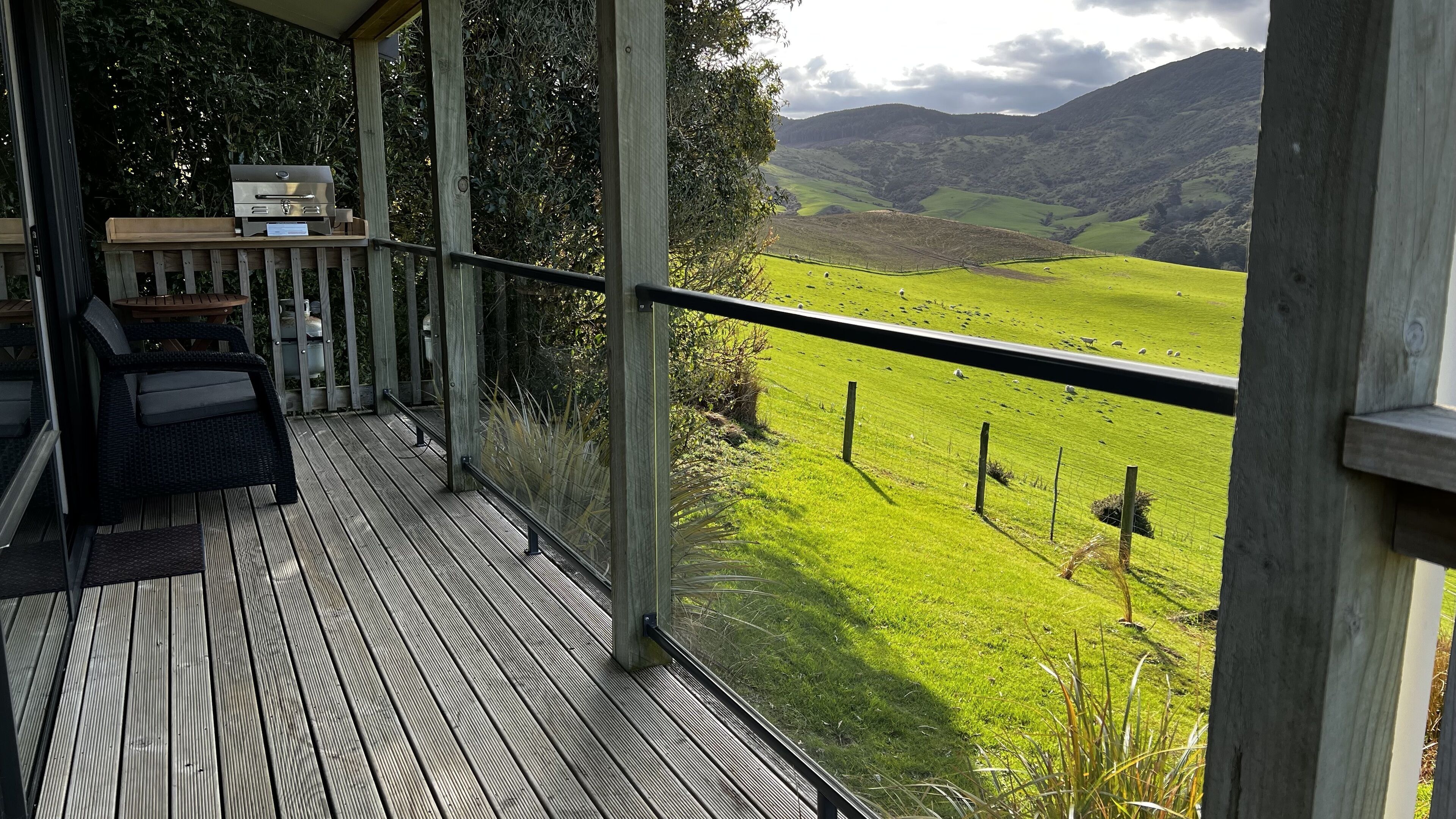 A wooden deck with a grill and seating area overlooking a green valley with sheep.