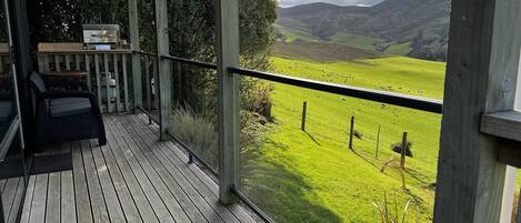 A wooden deck with a grill and seating area overlooking a green valley with sheep.