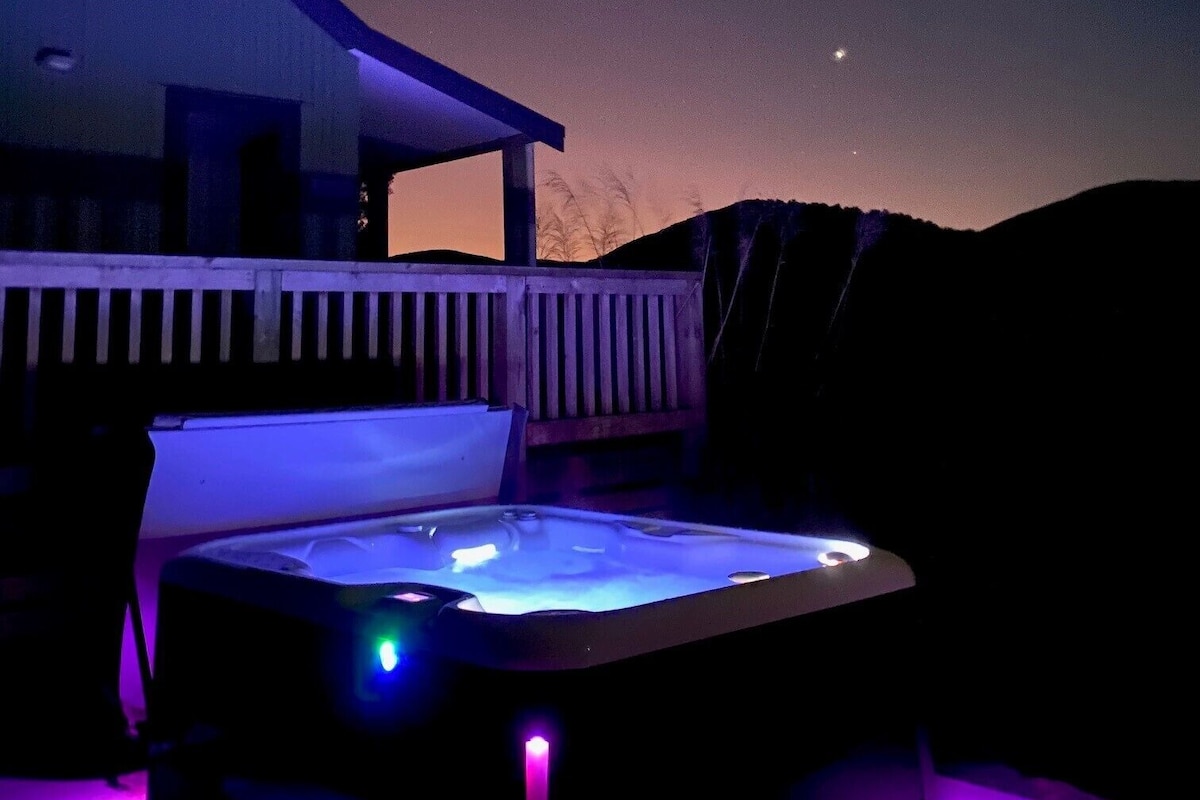 A hot tub with a view of the night sky, a wooden deck, and a building in the background.