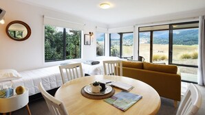 A room with a dining table, chairs, a sofa, and large windows offering a view of the countryside. - Mohua Park - Catlins Eco Accommodation (Tawanui)
