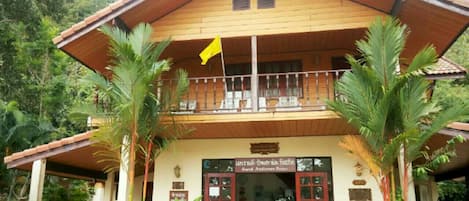 Un edificio de madera de dos pisos con un balcón, rodeado de vegetación tropical.