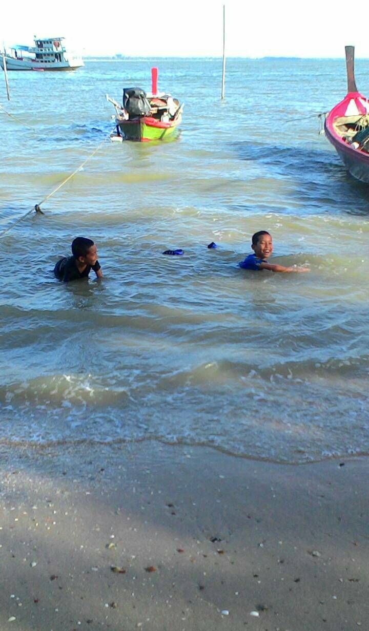 Deux personnes nagent dans une eau peu profonde, avec des bateaux en arrière-plan.