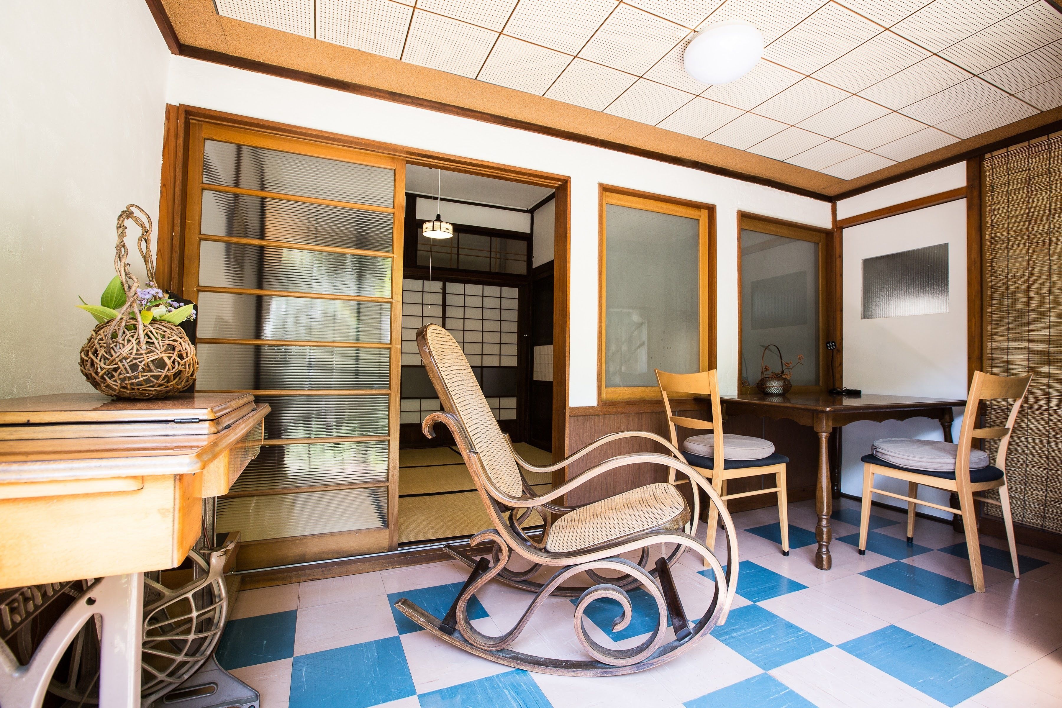 Una habitación japonesa tradicional con suelo de tatami, una mesa baja con una cesta tejida, un sillón mecedora y puertas correderas.