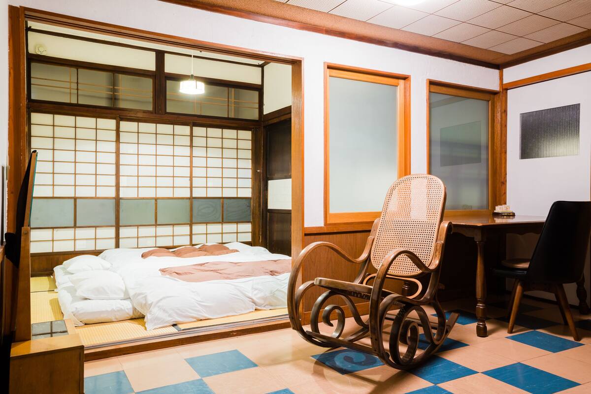 Ein traditioneller japanischer Raum mit Tatami-Bodenbelag, einem Futon, einem Holzschaukelstuhl und einem Schreibtisch mit Stuhl.