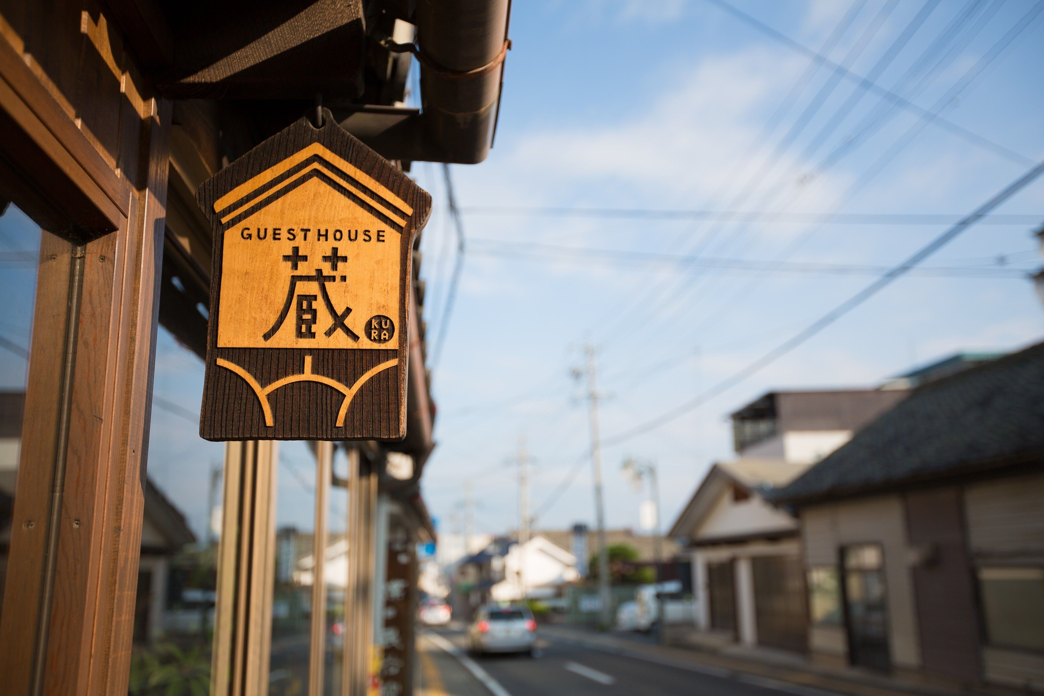 A wooden sign with "GUESTHOUSE" and Japanese characters, indicating a guesthouse on a street with buildings and power lines.