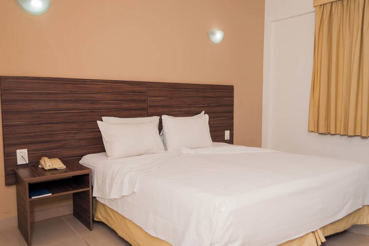 A neatly made bed with white linens, a wooden headboard, a bedside table with a telephone, and a wall-mounted light fixture.