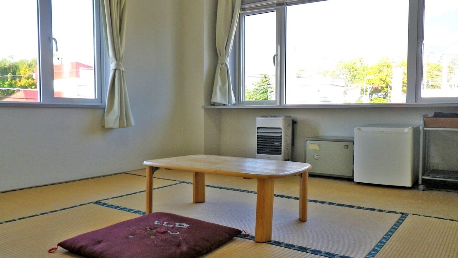 Une pièce avec un sol recouvert de tatamis, une petite table en bois, un coussin et une fenêtre avec des rideaux.