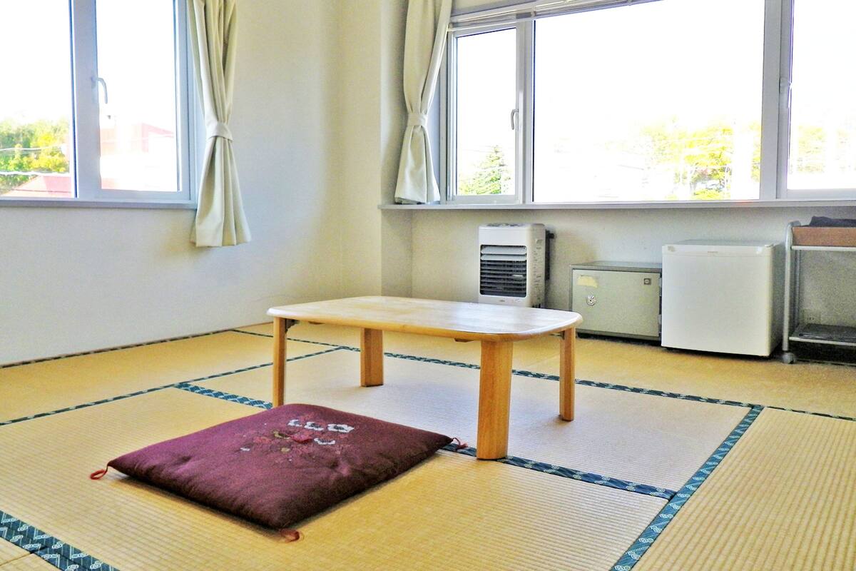 A room with tatami mat flooring, a small wooden table, and a cushion. There are two windows with curtains, a radiator, and a cabinet.