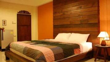 Cama de casal com colcha, cabeceira de madeira, mesa de cabeceira pequena com abajur e uma porta à esquerda.