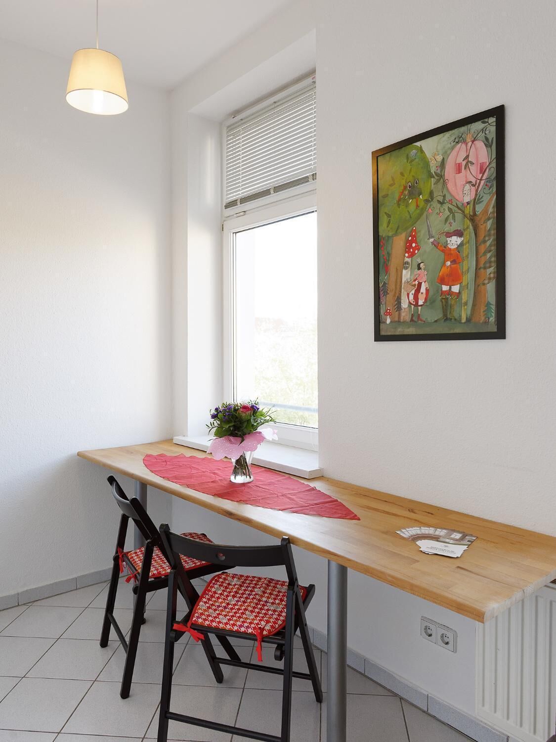 Un comedor con mesa de madera, dos sillas, una lámpara colgante, una ventana con persianas y un cuadro en la pared.