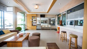 A modern hotel lobby with a wooden reception desk, a seating area with a sofa and ottomans, a bookshelf, and a large window with a view of the street. - Central place serviced apartment 1 (Chonburi)