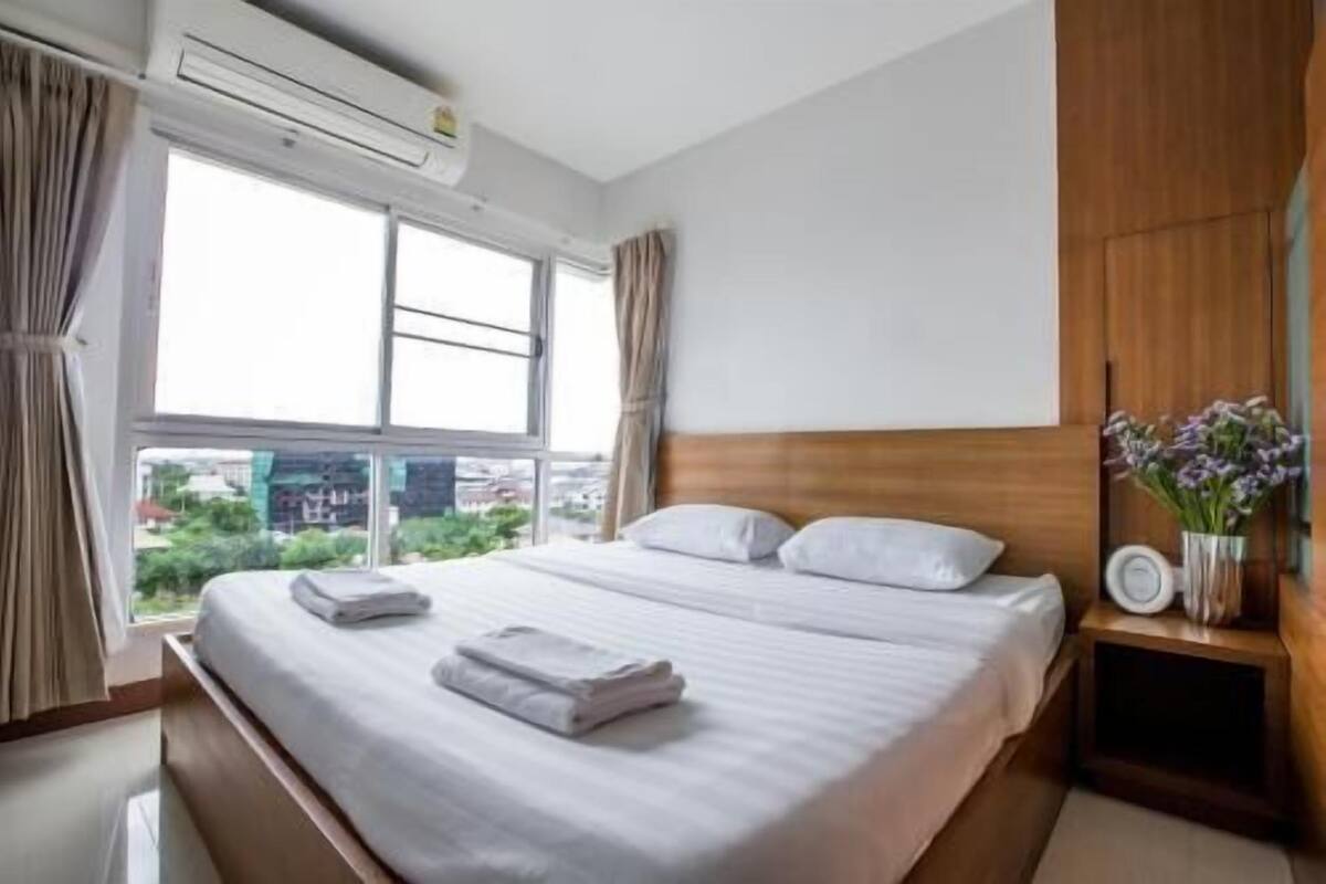Una habitación de hotel con una cama grande, un equipo de aire acondicionado, una mesita de noche con un jarrón de flores y una ventana con cortinas.