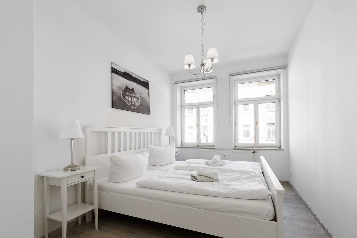 Une chambre à coucher blanche, avec un lit, des tables de chevet, un luminaire suspendu et une fenêtre.