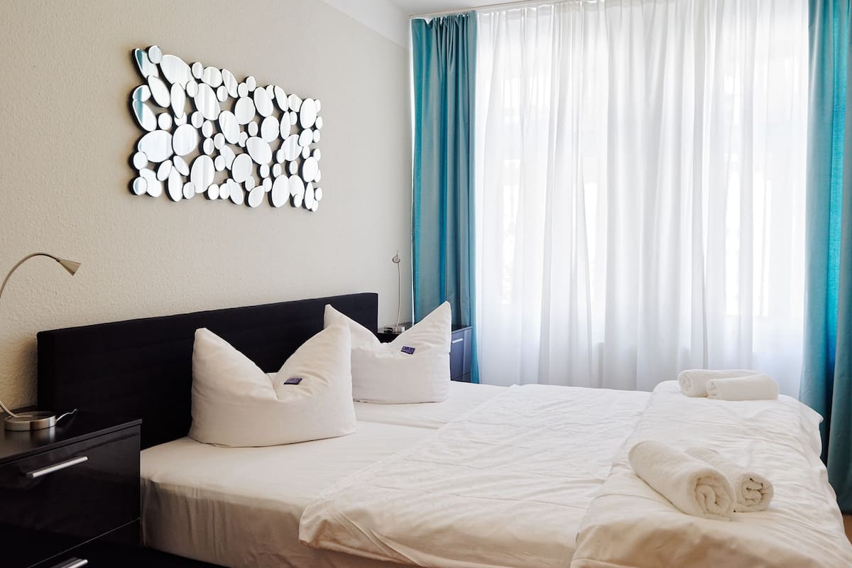 Une chambre d’hôtel avec un grand lit, du linge de lit blanc, une œuvre d’art moderne accrochée au mur et des rideaux de couleur turquoise.