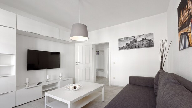 Un salon moderne avec un canapé blanc, une table basse blanche, un téléviseur à écran plat et un luminaire suspendu. Une porte donne accès à une autre pièce et un tableau encadré est accroché au mur.