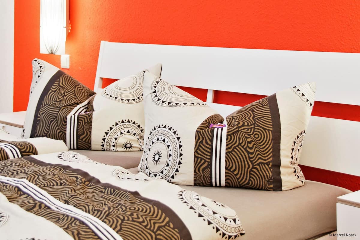 Un lit avec des coussins à motifs et une tête de lit blanche, dans une pièce aux murs orange.