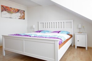 A bedroom with a white bed, a white nightstand, a colorful painting on the wall, and a sloped ceiling. - City Park Apartments Ludi (Leipzig)