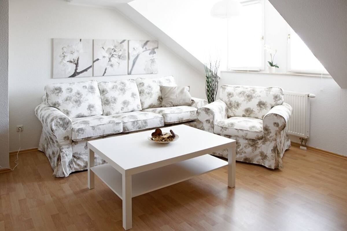 Un salon avec un ensemble de canapés blancs, une table basse blanche et un radiateur. Des œuvres d’art florales ornent le mur au-dessus du canapé, et une fenêtre offre une vue sur l’extérieur.