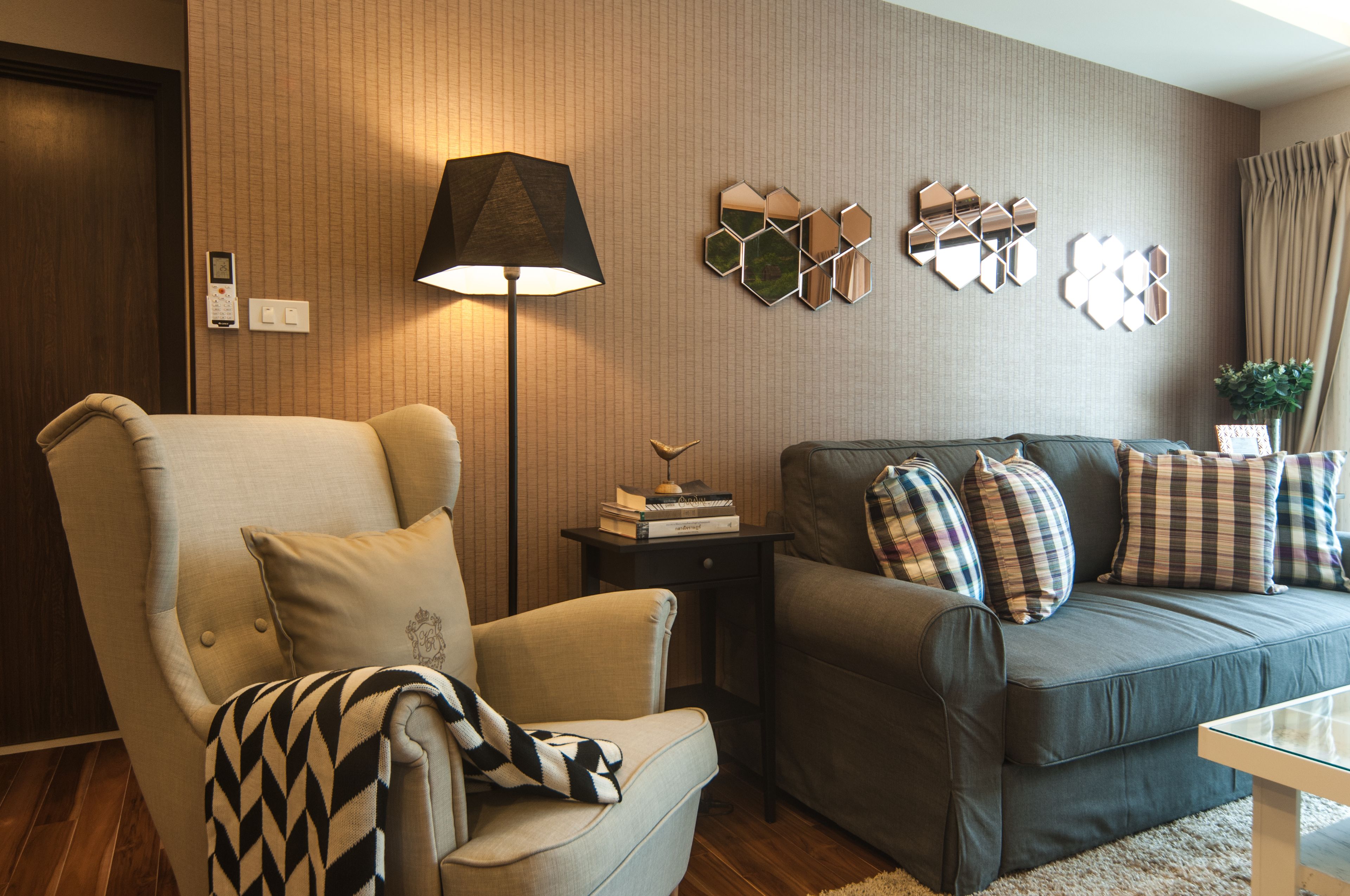 A living room with a sofa, armchair, and a lamp. There are hexagonal mirrors on the wall and a small table with books.