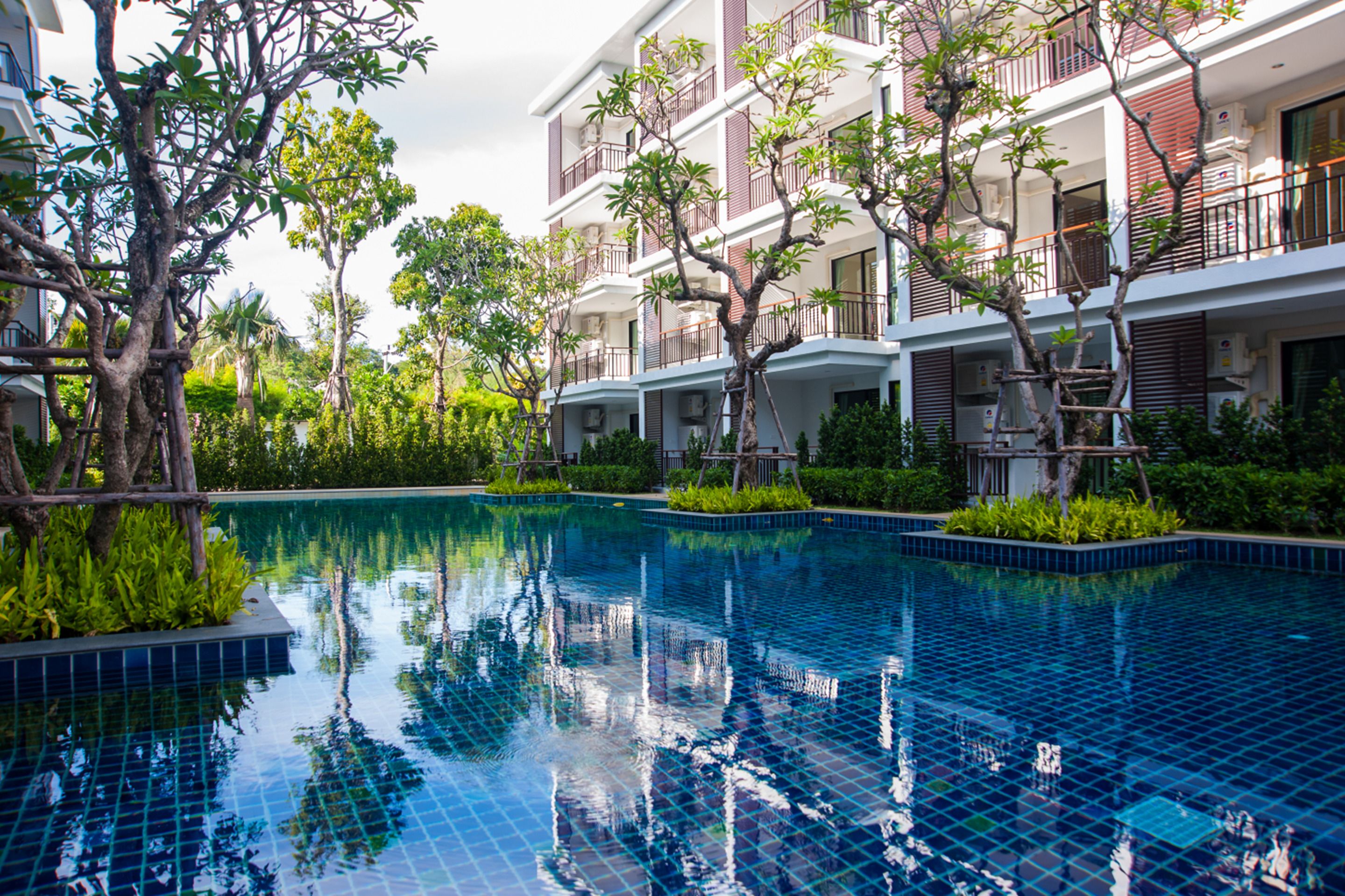 Piscina com bordas azulejadas, cercada por vegetação e um edifício com várias andares e sacadas.