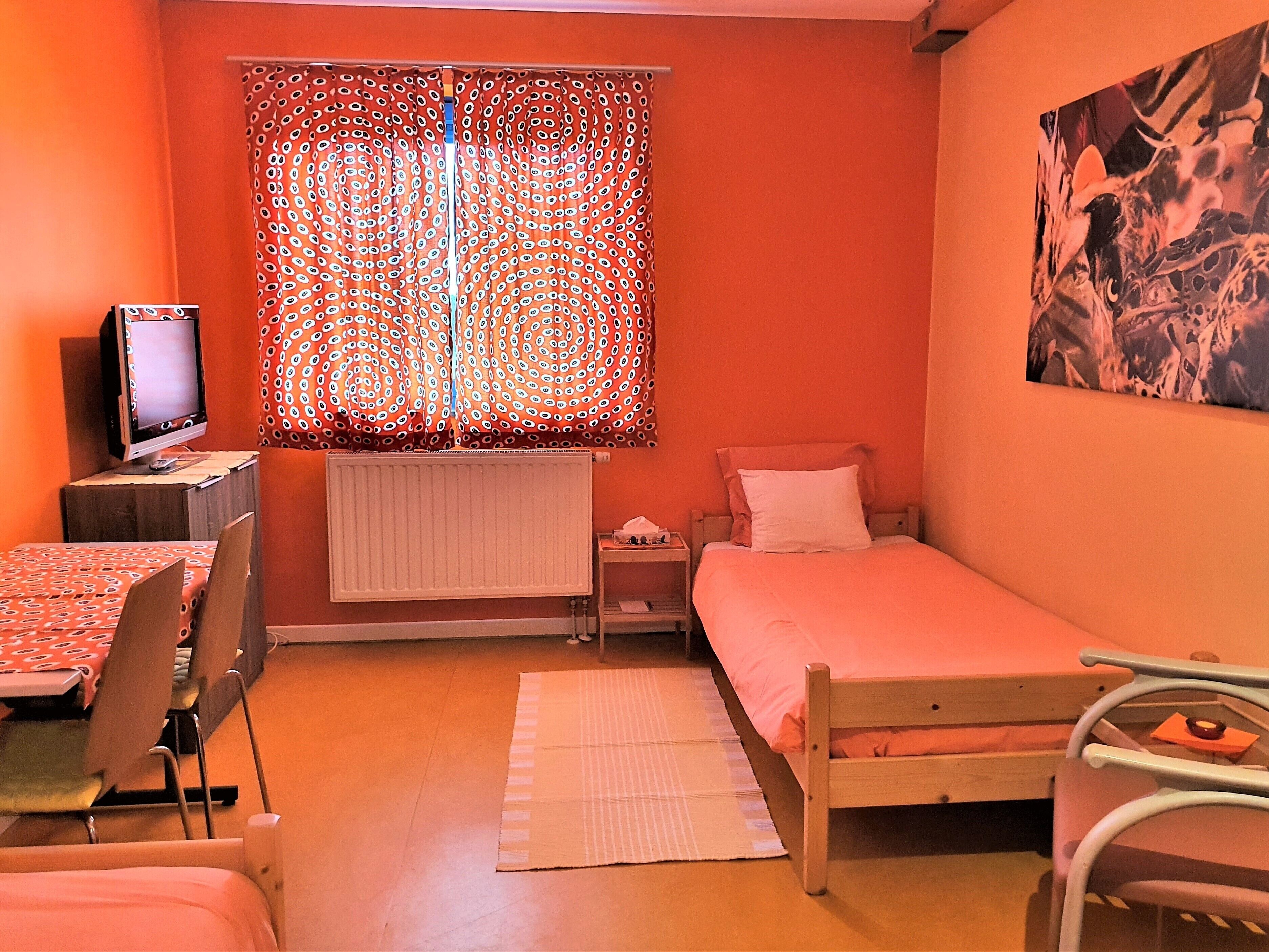 A small, orange-walled room with a single bed, a desk with a computer, and a window with patterned curtains.