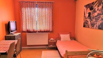 A small, orange-walled room with a single bed, a desk with a computer, and a window with patterned curtains.