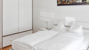 Un hotel moderno con un armario grande, una cama bien hecha con sábanas blancas, una mesita de noche con lámpara y un cuadro de un paisaje urbano en la pared.