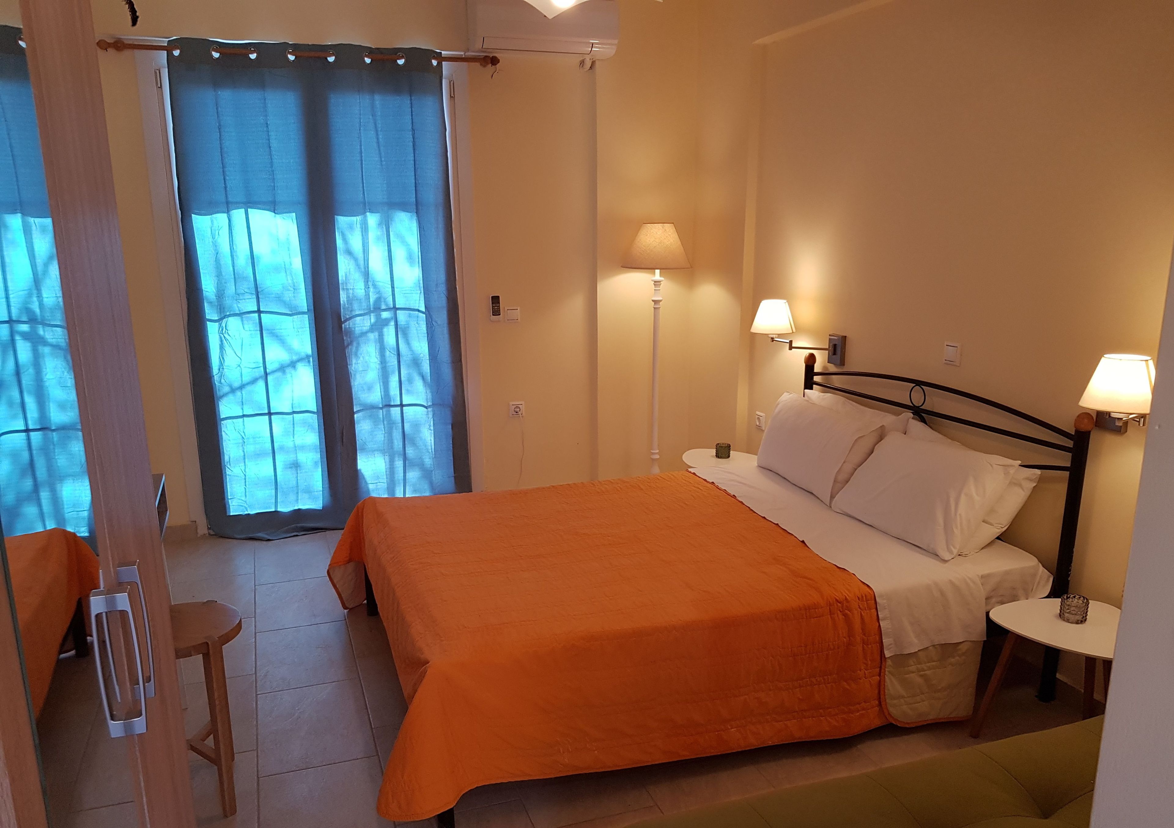 A bedroom with an orange bedspread, two bedside lamps, a small table, and a view through a window with curtains.