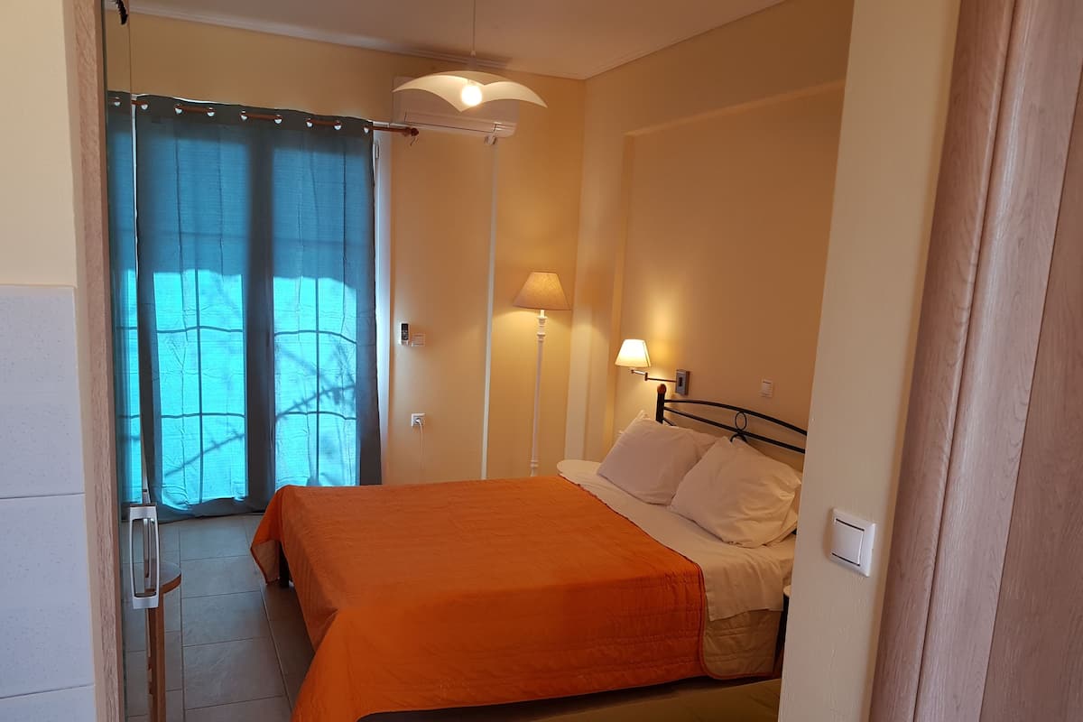Un dormitorio con colcha naranja, dos lámparas de noche y vistas a través de una ventana con cortinas translúcidas.