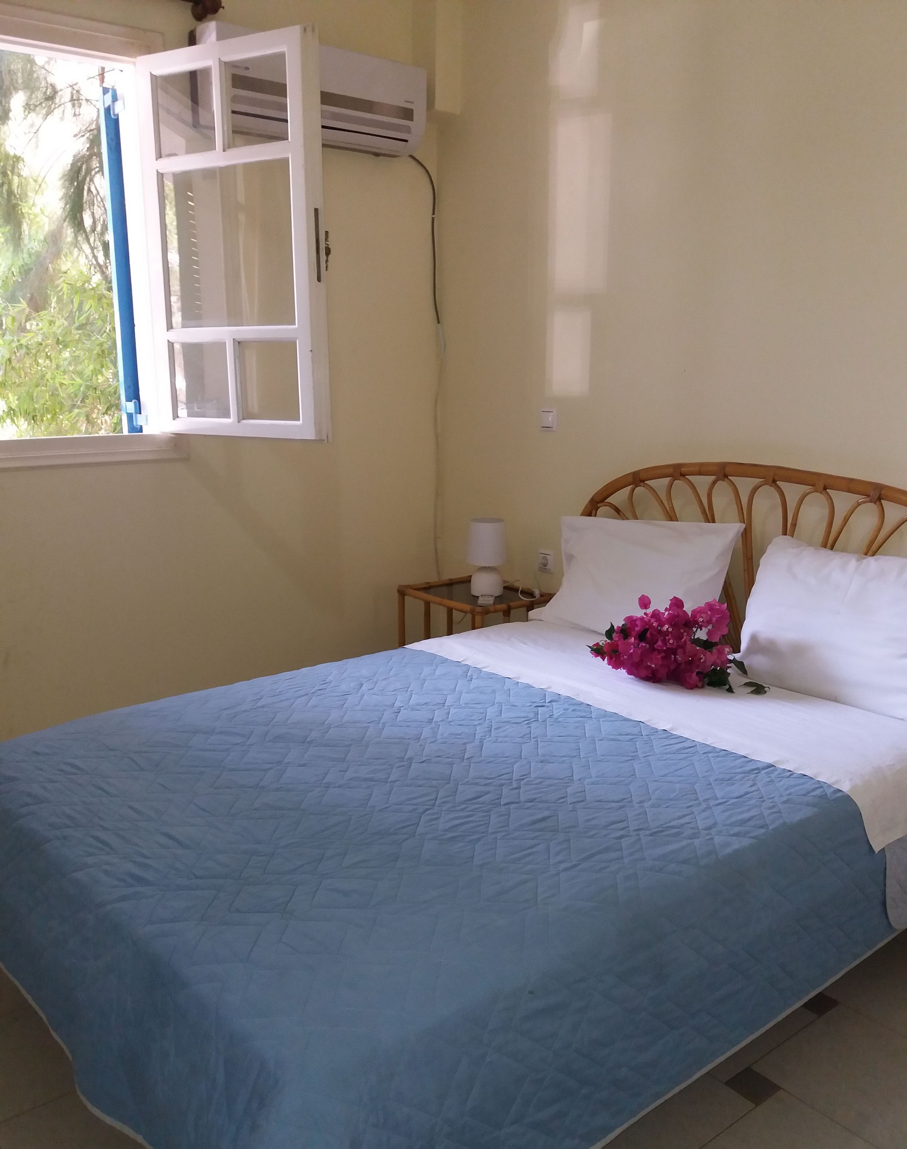 A bedroom with a bed, a nightstand, a lamp, and a window with an air conditioning unit.