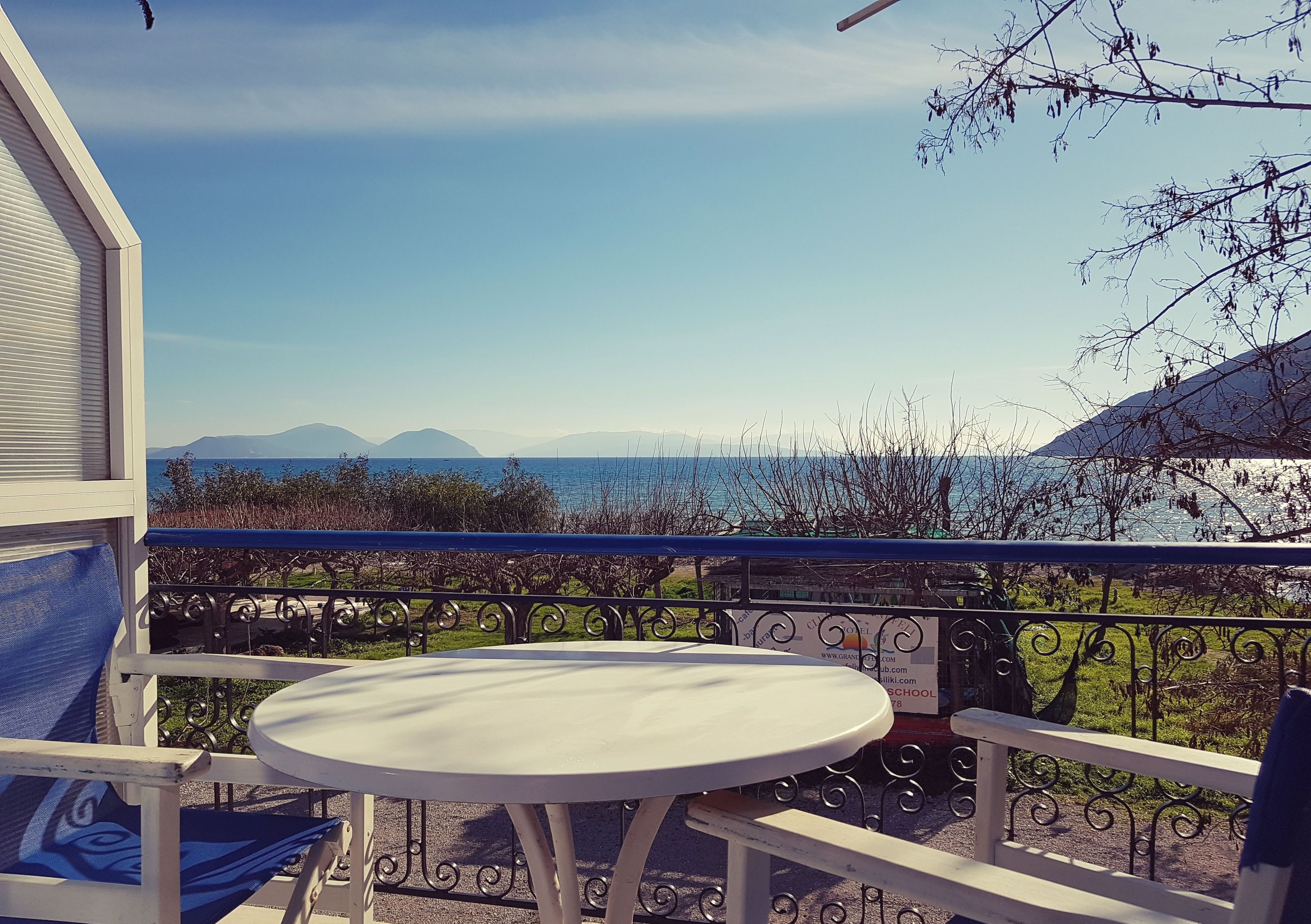 A balcony with a white table and blue chairs, overlooking a scenic view of the sea and mountains.