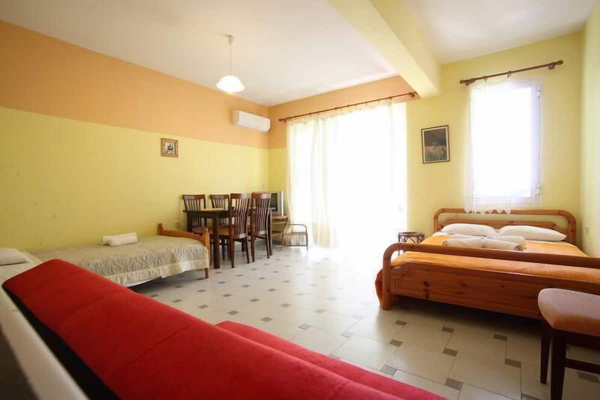 Una habitación con dos camas, una mesa de comedor con sillas, un sofá rojo y una ventana con cortinas.