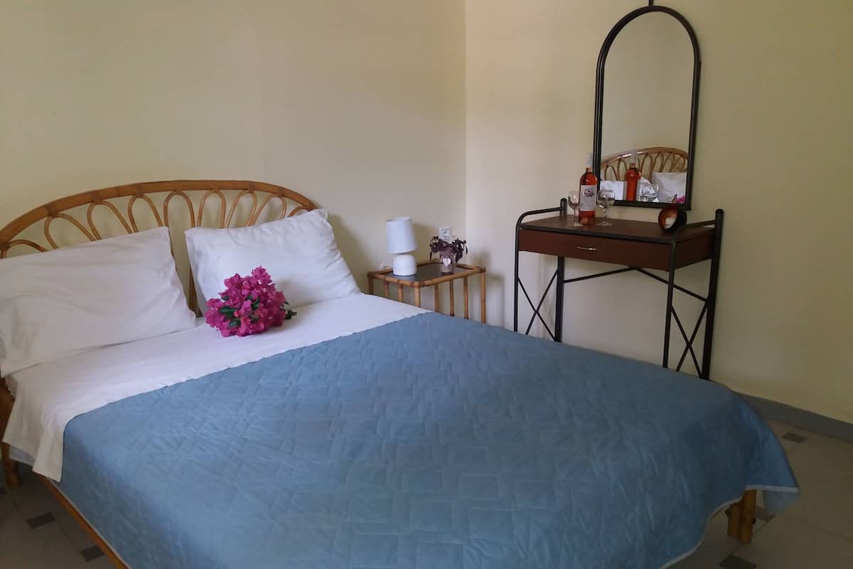 Una cama bien hecha con colcha azul, almohadas blancas y un arreglo floral rosa. Hay una mesita de noche pequeña con una lámpara y un espejo.