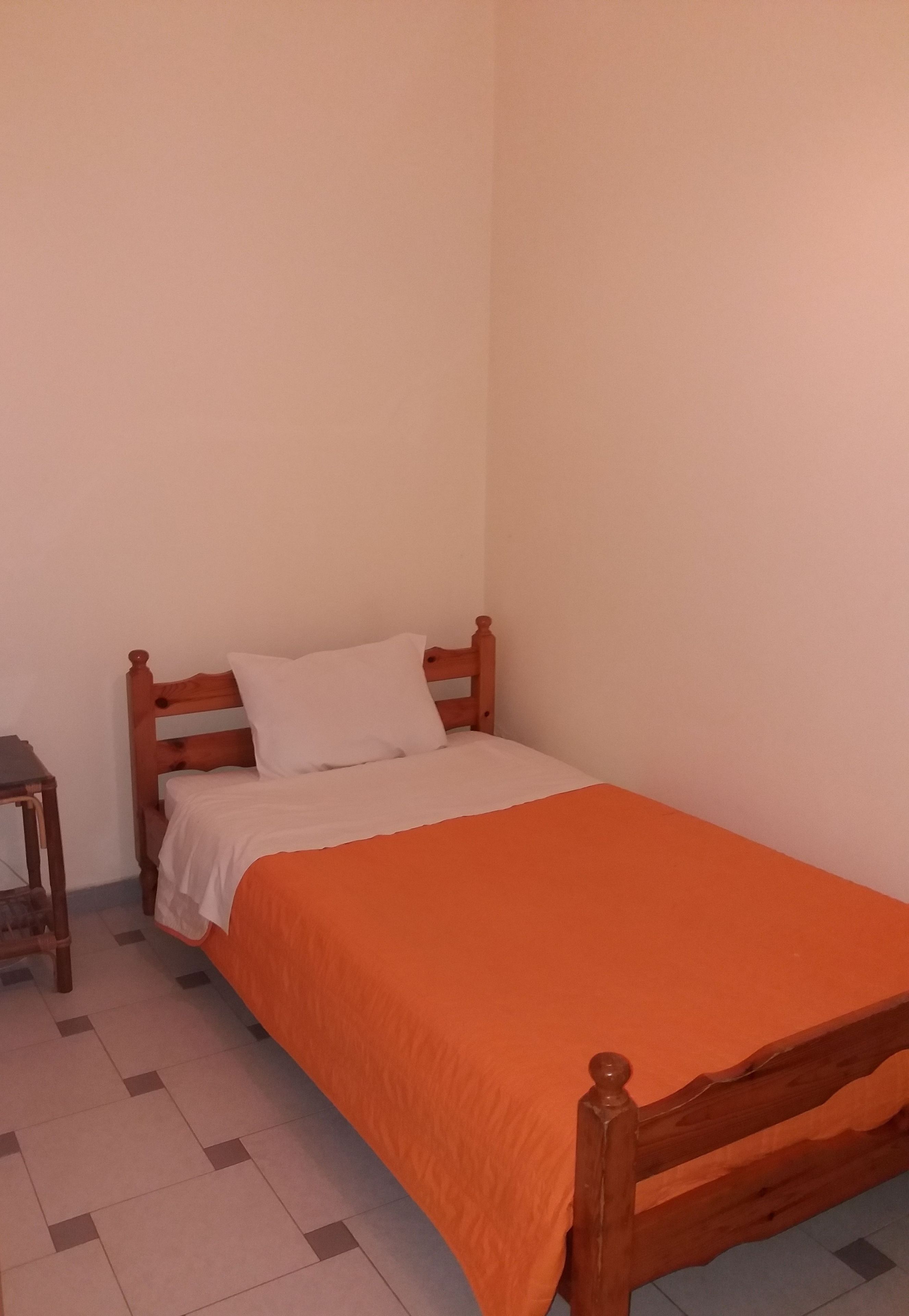 Un letto singolo con copriletto arancione e lenzuola bianche, una testiera in legno e un comodino in legno con una lampada.
