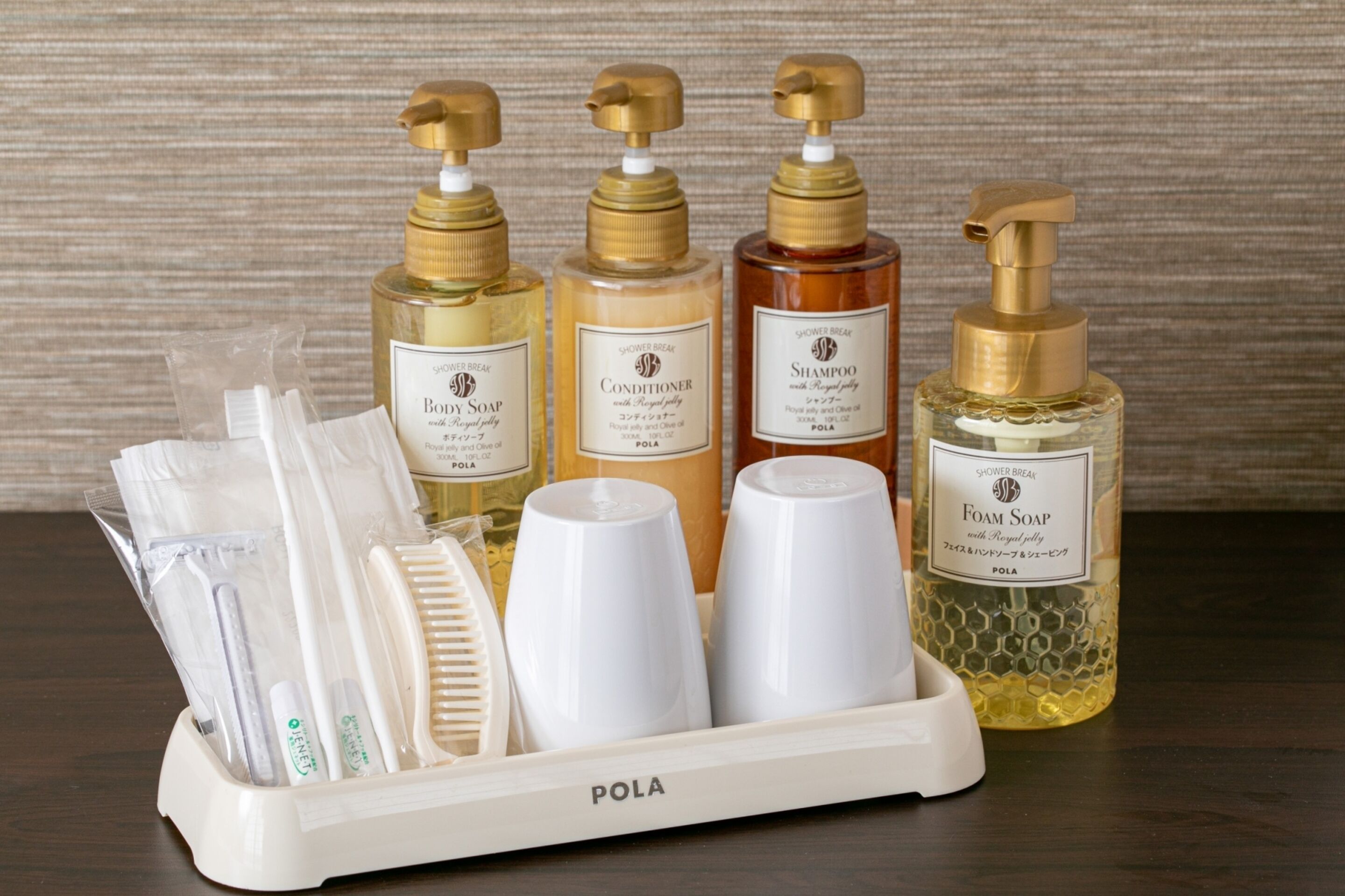 A set of toiletries including a bottle of body soap, conditioner, shampoo, and foam soap, along with several white cups and a hairbrush, all arranged in a white tray labeled "POLA".