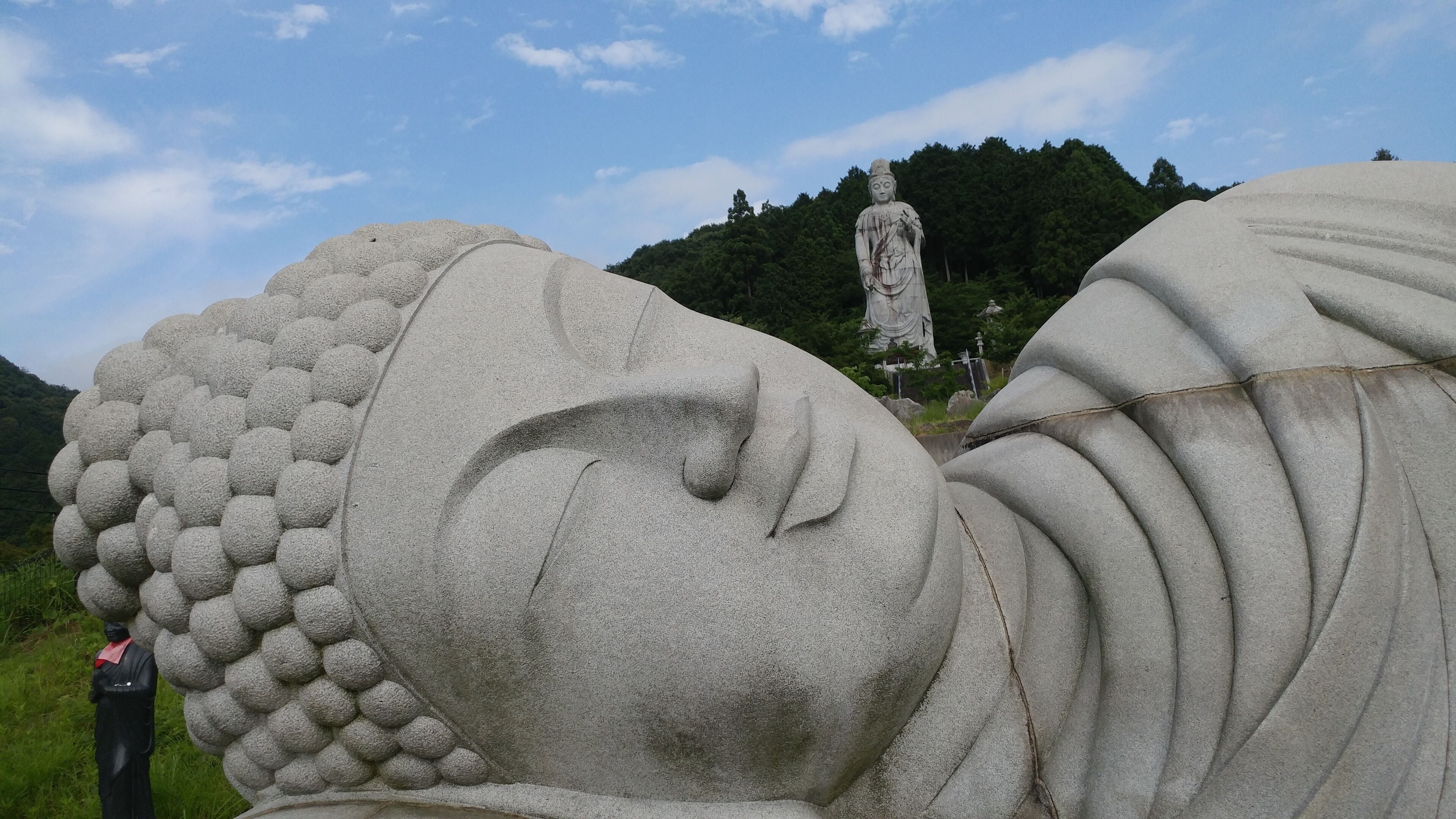 Une grande statue de pierre, dotée d’une tête aux détails soignés et d’une expression sereine, se dressant sur un fond de végétation luxuriante et d’un ciel dégagé.
